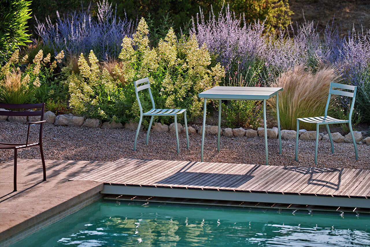 Set of light mint-coloured metal garden furniture by a table placed at the edge of a swimming pool, with ornamental plants in the background.