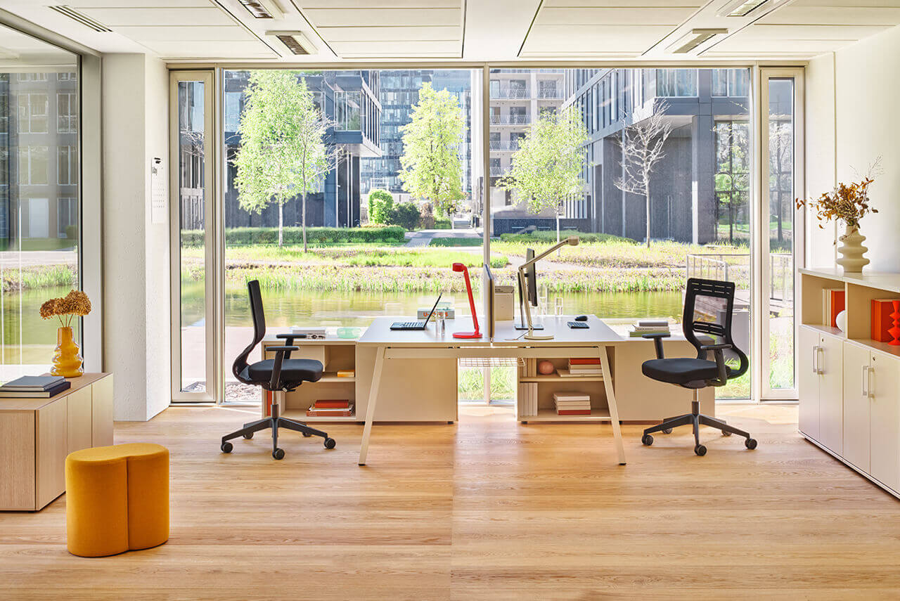 Modern office with two workstations, ergonomic chairs and a view of a green courtyard through large windows.