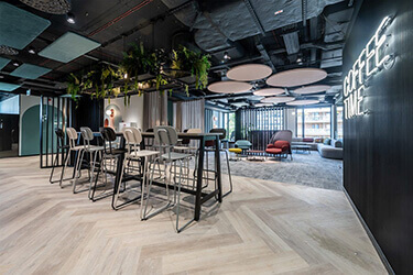 Office canteen equipped with modern high stools and high tables, plants and a neon sign ‘COFFEE TIME’ on a black wall.