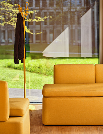 Yellow modular sofa set by a large window overlooking the greenery, next to a coat rack in a modern office interior.