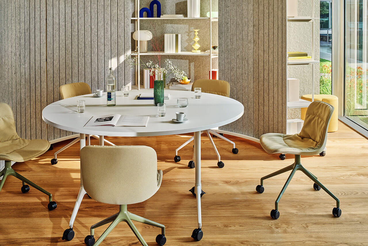Oval white Revi table surrounded by Baltic 2 chairs in a modern interior with shelving and large windows.