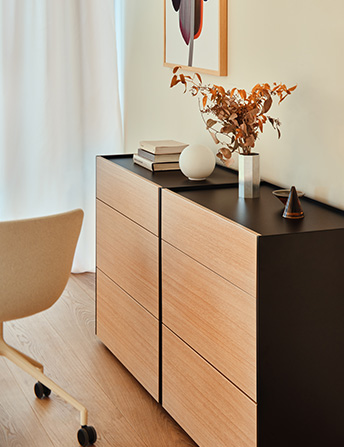 Minimalist office dresser in light wood and black top, set against a wall with ornaments and swivel chair.